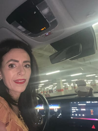 A woman with dark hair and red lipstick is in the driver's seat of a car. The car appears to be in an indoor parking structure, based on the overhead lighting. The rearview mirror and control panel are visible, as is a display screen showing "05:27 mar. 26-05" and "After You" playing, labeled with the name "ZAMB" beside it. There is also a teddy bear hanging on the windshield. The speedometer reads "5.6." The scene appears to be indoors, likely a parking garage or similar structure. The location is assumed to be in Mensdorf, Luxembourg based on the tags provided.