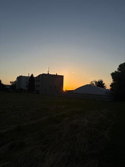This image captures a sunset scene in Tlustovousy, Czechia. The foreground is dominated by a dark, unkempt field of dry grass, suggesting a rural or undeveloped area within the town. In the mid-ground, a cluster of modern apartment buildings stands silhouetted against the vibrant orange and yellow hues of the setting sun. The buildings appear to be residential, with visible windows and balconies. To the right of the apartment buildings, a large, white, tent-like structure is visible, possibly a temporary event space or storage facility. A concrete fence separates this structure from the field. The sky transitions from a deep blue at the top to a warm gradient as it approaches the horizon, where the sun is setting behind the buildings. A faint contrail from an aircraft is visible in the sky. The overall atmosphere is serene, characteristic of the end of the day in a quiet suburban or semi-rural setting.