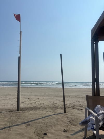 The image depicts a beach scene in Acapulco de Juárez, Mexico, under a clear, bright blue sky. In the left foreground, a tall, light-colored pole, possibly wood, is anchored in the sand. A red rectangular flag is affixed to its top, unfurled horizontally to the right, indicating wind presence. Several smaller horizontal protrusions are spaced along the pole's left side. A rope is tied near its base. To the right of this, a shorter, darker vertical pole is also embedded in the sand, casting a distinct shadow towards the right. In the far right foreground, a partial view of a multi-post structure, likely a beach cabana or shelter, is visible. Beneath this structure, a light-colored beach chair holds two folded towels: one predominantly white with blue accents, and another striped gray and white. The beach consists of light brown sand, showing numerous parallel tracks, consistent with vehicle activity, and scattered footprints. In the midground, the ocean extends to the horizon, featuring multiple breaking waves. A small, dark, humanoid figure is visible in the water beyond the immediate wave line. No other prominent individuals are present. Strong sunlight is indicated by the clear shadows cast by the poles.