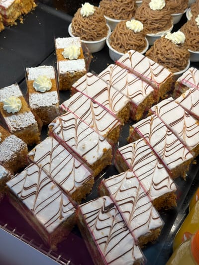 The scene displays a commercial display case filled with assorted pastries. In the upper right, approximately fifteen individual Mont Blanc pastries are presented in white paper cups, each topped with a light-colored cream swirl over a mounded brown chestnut cream piped in a noodle-like pattern. The central and lower portions of the display are occupied by multiple rows of rectangular slices of cream cake or mille-feuille, featuring white icing with wavy brown decorative lines. Many of these slices are individually separated by clear plastic film, with some groups secured by thin pink plastic banding. To the upper and middle left, several rectangular multi-layered cake slices are visible, dusted with powdered sugar and topped with a small dollop of white cream. The pastries are arranged on a dark, flat surface, with some in the foreground resting on a dark metal grate. Partially visible in the bottom right are containers of yellow liquid, indicating beverages. No individuals are present. The setting is a retail bakery or patisserie display in Fribourg - Freiburg, Switzerland; a specific location within the city is not identifiable from the image.