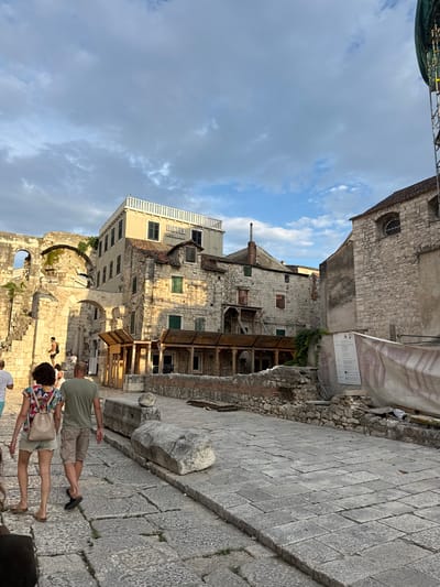 This media file depicts an outdoor scene in Split, Croatia. The image features ancient stone buildings with visible signs of age and some areas of reconstruction or preservation, evidenced by scaffolding and temporary structures. The architecture appears historic, with stone blocks and arched openings suggesting Roman or similar ancient origins.
In the foreground, several people are walking on a stone-paved walkway. A couple is prominent, with the man wearing a green t-shirt and beige shorts, and the woman in a colorful patterned shirt and denim shorts, carrying a light-colored bag. Other individuals are partially visible further back. The overall atmosphere suggests a tourist or pedestrian area within a historical site.
The sky is partly cloudy, with a mix of blue sky and white clouds, indicating daytime. The lighting suggests it might be late afternoon or early evening, given the angle of the sun and the shadows. The setting is clearly an urban environment characterized by historical architecture.
One sign is visible on a banner attached to a building, displaying some text and graphics, though it is not legible in detail. Another sign with text is attached to a wooden structure. The overall impression is one of exploring historical ruins or ancient city streets.