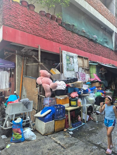The image depicts a narrow street scene in Parañaque, Philippines, crowded with discarded and stored items. A young girl in a blue shirt and shorts is walking along the street, holding a lollipop.  Numerous objects are piled up along the side of the building, including plastic crates, storage containers, a pink teddy bear, and various covered items. Political posters with the names Edwin Olivarez and Dra. Raquel Gabriel are affixed to the building. The building has a red roof and walls. A green netting is visible above. The street surface appears wet and uneven.