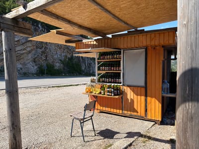 This media file depicts a small, open-air roadside stand selling bottled beverages and jars of preserves. The stand is constructed from orange corrugated metal and features a wooden shelter with a roof made of oriented strand board (OSB). Several shelves are visible, stocked with numerous green glass bottles and an assortment of glass jars. In front of the stand, a shelf displays more bottles and some fruits, including what appear to be oranges and bananas. A single, rustic metal chair with a patterned cushion sits in the gravel in front of the stand.
The scene is set against a backdrop of a rocky hillside and a paved road, suggesting a location along a scenic route. The time of day is indicated by the strong shadows cast by the stand and the chair, suggesting it is a sunny afternoon. The weather appears to be clear and bright, with a blue sky visible. The overall atmosphere is one of rural commerce, likely catering to travelers. No people are visible in the image. There is no discernible text visible in the media file. The location is identified as Ponikvica, Montenegro.
