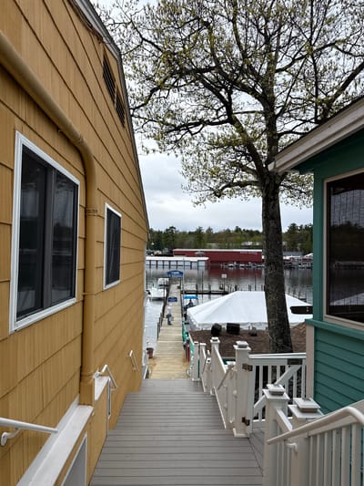 The image depicts a partially obscured view of Paugus Bay on Lake Winnipesaukee, in Laconia, New Hampshire, United States. A yellow-painted building with two windows and a visible vent occupies the lower portion of the frame. Above this, a tree with nascent leaves stretches towards the left, its branches partially overlapping a light blue building. The center reveals a wooden walkway leading towards the bay. Boats are visible on the water, along with covered docks and buildings on the opposite shoreline. The sky is overcast.