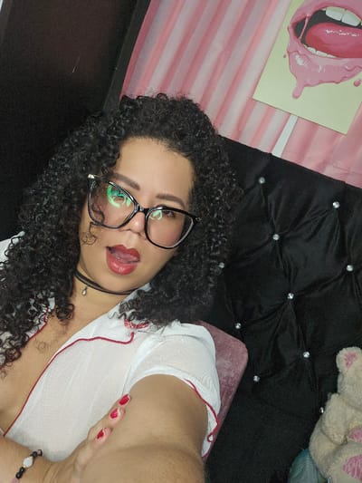 A woman with dark, curly hair and black-framed glasses is captured in a selfie, angled diagonally from the upper right to the lower left. She gazes directly at the camera with a playful expression, her tongue slightly extended past her red-lipsticked lips. She wears a white, sheer top with red trim and a black choker necklace featuring a small, teardrop-shaped pendant. Red nail polish is visible on her fingernails, and a multi-colored beaded bracelet adorns her wrist.
The background features a black, tufted headboard or cushion embellished with several sparkling, button-like studs. Behind her head and to the left, a pink and white striped pattern is visible, along with a piece of wall art depicting an open mouth with pink lips, white teeth, and a pink tongue with dripping liquid. A light-colored, possibly plush toy is partially visible in the upper right corner of the image. The setting appears to be an indoor space, likely a bedroom, within Perímetro Urbano Cúcuta, Colombia.