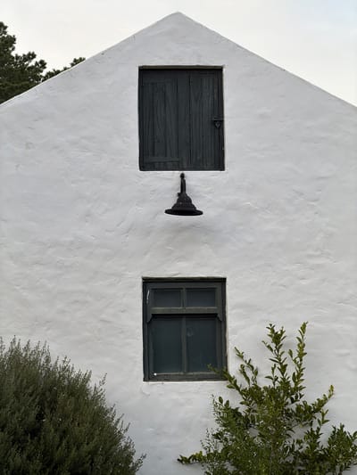 The exterior of a building in the Overstrand Local Municipality, South Africa, features a textured white wall. There are two dark-framed windows, one closed with a solid wooden shutter secured with a latch, and another open with multiple panes. A black, conical outdoor light fixture is mounted between the two windows. Green foliage, including branches and a bush, partially obscures the right window and the lower right corner of the building. The sky is visible in the lower left corner.