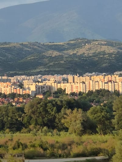 This image presents a panoramic, elevated view of an urban landscape in Moshtanets, Bulgaria, under warm, low-angle sunlight, indicative of either early morning or late afternoon.

In the far background, a prominent range of hazy, blue-grey mountains spans the horizon, displaying gradual slopes and muted tones due to atmospheric perspective. Closer to the viewer, a series of rolling hills are visible, covered in a mix of sparse, dry, golden-brown vegetation and darker green patches of trees. A few indistinct, small structures are discernable on the crests of these hills.

The midground is dominated by a densely populated urban area, characterized by numerous multi-story apartment buildings. These structures primarily feature facades in shades of beige, cream, and warm peach or orange, brightly illuminated by the directional sunlight, which casts distinct shadows on their sides and between buildings. The architectural style appears functional and consistent across the visible residential blocks.

Immediately in front of the urban area, a thick band of lush, dark green deciduous trees forms a natural boundary. Below this tree line, in the foreground, is an area of mixed vegetation, including brighter green shrubs and significant patches of reddish-brown or ochre-colored foliage, possibly dry grass or specific plant types. The very bottom of the frame shows an unpaved, uneven surface, likely a track or road, with a partial, weathered concrete or stone structure visible on the far left.

No individuals, animals, or explicit actions are discernible within the frame. The scene conveys a sense of quietude, with the city nestled between natural, mountainous terrain.