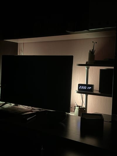 The image depicts a dimly lit room, likely a bedroom or office, at night. A large, black television screen is prominently positioned on a dark wooden desk in the foreground. To the right of the television, on a shelf, a small digital clock displays "23:30" and "23°C". A black speaker is also visible on the same shelf. In the background, a pink, textured wallpaper covers the wall. A desk organizer with pens and pencils is on another shelf above the clock. The overall impression is one of a quiet, private space. There is no indication of the specific location being in Bryansk, Russia, or any other city.