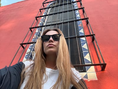 A fair-skinned blonde wearing sunglasses and a white top is shown upside down in front of a red wall with a window. The window has blue and white patterned tiles around it and a metal security grate. Her hair falls past her shoulders, and a dark-colored jacket is partially visible on the upper right. The perspective gives a distorted view.