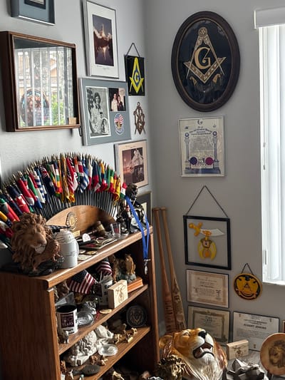 The image shows a collection of masonic and other memorabilia displayed on shelves and walls in a room, possibly a home office or den. The items include framed certificates, photographs, flags, and decorative objects such as lion statues. The room appears to be well-lit, with natural light coming from a window on the right side of the frame.

Specifically, on the wall, there is a large framed Masonic Square and Compasses emblem with a "G" in the center. Below it are several framed documents and photographs, including what appears to be a certificate of membership and a photo of an astronaut. To the left, a display case holds a collection of what look like small flags or banners. A fan-shaped display of various national flags is positioned on a shelf.

On the shelves below, more decorative items and memorabilia are arranged. These include more lion statues, a baseball, a gavel, a small American flag, and several other framed items. The overall impression is of a personal collection, carefully curated and displayed.

The image is geolocated to Sun City Center, Florida, United States of America.