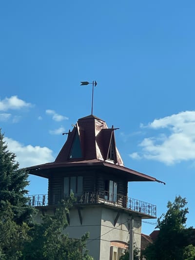 A weather vane shaped like an arrow sits atop a conical, dark red roof. The roof extends outward, forming an overhang. Below the roof is a wooden structure with two tall windows. This structure is supported by a lighter colored, cylindrical base, which is surrounded by a balcony with a decorative railing. The balcony is also partially obscured by lush green trees. The background is a clear blue sky with scattered white clouds.