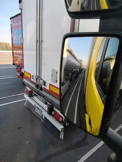 The image shows a close-up view of the side and rear of a white semi-trailer truck. The truck has "www.sealane.c" and "SEALAN" printed on a red panel on its side, along with a European Union flag motif. Below this, "INTERNATIONAL TRANSPORT", "+44 (0) 1582 40", and "sales@sealane.c" are visible. The rear of the truck shows a license plate "LF-27 SKN" and the text "SCHMITZ CARGOBULL" with "The Trailer Company." The image is taken from the perspective of a vehicle, likely another truck, as a side mirror reflects other vehicles, including a yellow truck cab and several tanker trucks lined up. The environment appears to be an outdoor lot or parking area with asphalt pavement marked with white lines. The background suggests an industrial or port area, with some fencing and structures visible. The scene is set in the city of Loon-Plage, France.