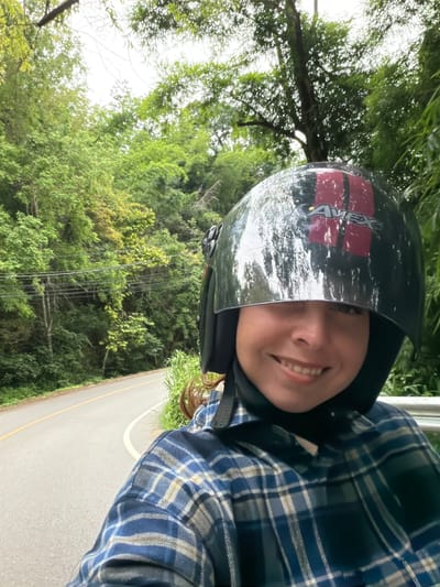 A person is on a motorcycle on a road in Samoeng Tai, Thailand. The person is wearing a helmet and a blue plaid shirt. The road is paved and has a yellow line down the middle. The surrounding area is a lush, green forest with tall trees. The sky is overcast.
