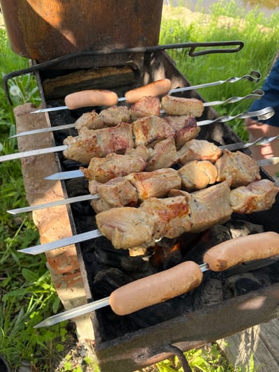 In Маяк, Russia, a close-up shot captures a barbecue with skewers of meat and sausages cooking over charcoal. The scene is set outdoors, with lush green grass and a body of water visible in the background, suggesting a natural or park-like setting. The barbecue grill itself appears to be made of metal and brick, with remnants of previous fires evident in the charred coals. The meat, cut into cubes, is skewered and shows signs of being grilled, with some pieces lightly browned and others more deeply seared. Several sausages, also on skewers, are positioned near the meat, indicating a mixed grill. The overall atmosphere is casual and suggests a leisurely outdoor meal or gathering.