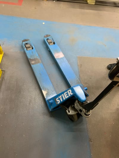 A blue pallet jack, labeled "STIER" with a maximum load capacity of 2500kg, rests on a blue and grey tiled floor in what appears to be a warehouse or industrial setting. The pallet jack is positioned at an angle, with its forks extended. One of its handles is visible, along with a person's black shoe and a small portion of their leg wearing dark pants. The surrounding environment is a hard, flat surface, possibly concrete or industrial flooring, with some debris scattered about. The lighting suggests an indoor environment, possibly with artificial overhead lighting. The scene is static, with no apparent activity beyond the presence of the pallet jack and the partially visible person.