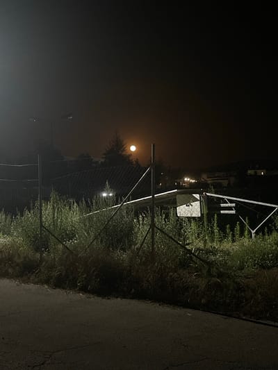 The image depicts a dimly lit outdoor scene at night. In the foreground, a chain-link fence runs horizontally across the frame, partially obscured by tall, wild vegetation. Beyond the fence, there is an area with some kind of structure or equipment, including what appears to be a white panel with a grid pattern. Further in the background, a full moon is visible in a hazy, dark sky. Several distant lights, possibly from buildings or streetlights, are also present. A single lamppost is visible on the left side of the image, its light partially illuminating the foggy atmosphere. The overall setting suggests an unkempt or industrial area on the outskirts of a town, possibly Vedrare, Bulgaria, given the context of the prompt. There are no people or discernible actions taking place in the image.