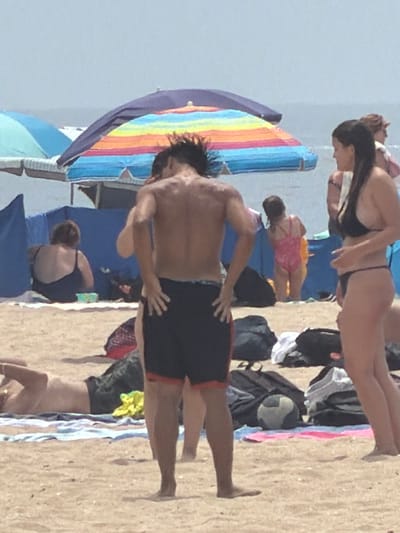 An outdoor, eye-level medium shot captures a beach scene in Espinho, Portugal, under a bright, hazy sky. In the foreground, a man with dark hair and a muscular physique, wearing dark swim trunks, stands with his back to the camera, facing the ocean. His left arm is bent at the elbow, and his left hand is resting on his hip. To his right, a woman with long dark hair, wearing a black bikini, is partially visible from the side. Her head is turned away from the camera, and her hands are clasped in front of her. In the background, a person in a pink swimsuit is visible. Further back, another individual, viewed from the back and wearing a dark swimsuit, sits on the sand. Two beach umbrellas are prominent; one is a large rainbow-colored umbrella, and another is a solid blue umbrella. Bags and towels are scattered on the sand, and a patterned beach ball rests on a blanket. The ground is composed of sand, and the ocean is visible in the distance. The overall atmosphere is that of a typical summer day at the beach.