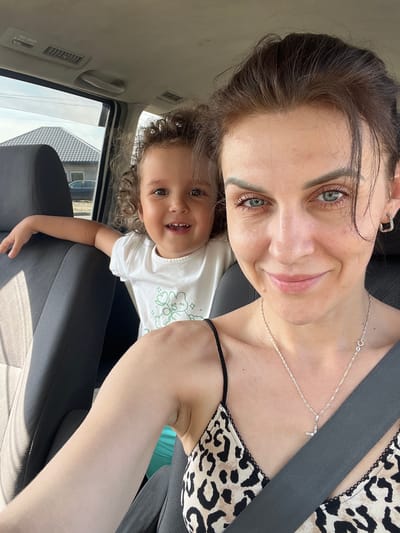 A woman and a young child are inside a vehicle. The woman is in the foreground, taking a selfie. She has brown hair, blue eyes, and is wearing a leopard print top and a silver necklace with a pendant. The child, with curly brown hair and brown eyes, is to her left, smiling and wearing a white t-shirt. They are seated in the car, which appears to be parked or moving slowly, with a view of houses and trees through the windows. The overall scene suggests a casual outing or travel. The image was taken in Aksu, Kazakhstan.