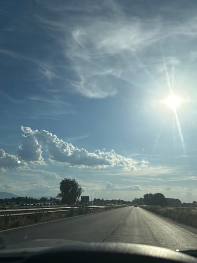 A drone's-eye view captures a sun-drenched highway leading into Gorni Bogrov, Bulgaria. The sun, a bright orb in the upper right, casts long shadows across the asphalt and illuminates the wispy cirrus clouds scattered across the brilliant blue sky. In the distance, a lone car travels south on the highway, its form a small silhouette against the vast landscape. To the left, a line of parked cars suggests a roadside attraction or rest stop, bordered by a metal guardrail. A solitary, windswept tree stands sentinel beside the road, its branches reaching towards the sky. The right side of the highway is flanked by a field of dry, golden grass, hinting at the summer season. In the far distance, rolling hills, shrouded in a haze, form the backdrop to this serene rural scene.