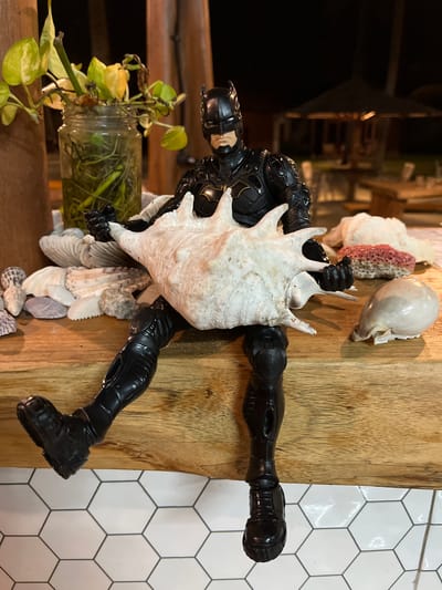 A Batman action figure is seated on a wooden surface, holding a large conch shell. The action figure is black with gold accents on its chest. It is positioned on a wooden ledge, with its legs dangling over the edge. Several seashells, coral, and a glass jar with a plant are arranged around the action figure on the ledge. In the background, there is an outdoor seating area with tables and umbrellas, suggesting a restaurant or resort setting. The ground below the ledge is tiled with a hexagonal pattern. The scene appears to be set at night, with artificial lighting illuminating the area. The location is identified as Legonipah, Indonesia.