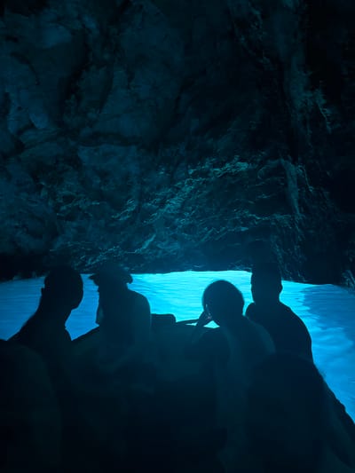 This image shows a group of people in a boat inside the Blue Cave, a sea cave on the island of Vis, near the town of Komiža, Croatia. The cave's interior is illuminated by a surreal, bright blue light, created by sunlight reflecting off the sandy seabed and passing through an underwater opening. The people are silhouetted against the glowing water, appearing as dark shapes within the dimly lit cave. The rocky walls of the cave rise steeply above the water, their rough textures visible in the faint light. The overall scene conveys a sense of mystery and wonder, with the vibrant blue water contrasting dramatically with the dark cave and its occupants.