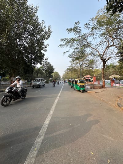 The image captures a daytime street scene in Vadodara, India, depicting a multi-lane asphalt road under a clear light blue sky. A faded white lane divider separates the road. On the left, a person wearing a white shirt and a light-colored helmet rides a black motorcycle, moving from left to right across the foreground. Behind the motorcycle, a silver van proceeds in the same direction, followed by additional distant motorcycles. On the right side of the road, a line of at least five green and yellow auto-rickshaws are parked parallel to the curb. Adjacent to these vehicles, a paved area features several small, reddish-brown structures resembling vendor stalls, one with a yellow awning, and another with a white umbrella covering a partially visible individual in a blue shirt. A low red brick wall or fence runs along this right-hand side, displaying partially visible signage. Dense green trees line both sides of the street, casting long shadows diagonally across the road from a light source to the left.