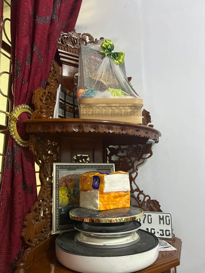 The image shows an interior scene, likely a home in Terang-terang, Indonesia. On a wooden shelf, there is a decorative basket wrapped in netting and tied with a green bow. Inside the basket, a book with "AL-QUR'AN" printed on it is visible, along with some artificial flowers. Below the shelf, a framed photograph of a family is displayed. Stacked on top of each other are two packages of tissues, one white and the other orange. A white and black electric appliance, possibly a stove or a fan, is placed underneath the tissues. To the right of the appliance, a license plate with the text "870 MG 10-3G" is leaning against the wall. A dark red curtain with a patterned design hangs to the left of the shelf. The overall environment appears to be cluttered with various decorative items and everyday objects.