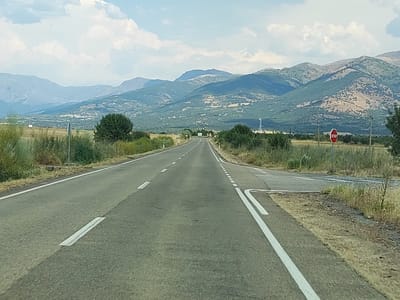 This media file shows a scenic drive through a rural landscape. The foreground features an asphalt road with clear white lane markings, suggesting it's a well-maintained route. The road appears to be leading towards a range of mountains in the distance. To the right of the road, a red octagonal STOP sign is visible, indicating a potential intersection or hazard ahead.

The landscape on either side of the road is dry and covered with sparse vegetation, consistent with a warm climate. Tall grasses and bushes line the roadside, with some scattered trees. In the mid-ground, more trees and shrubs are visible, and beyond that, the foothills of the mountains are covered in a mix of vegetation and possibly some cultivated land. A small village or settlement can be seen nestled in the valley between the foothills and the mountains.

The mountains themselves are rugged and imposing, with varying shades of green and brown indicating different types of terrain and vegetation. The sky is partly cloudy, with soft white clouds against a pale blue backdrop, suggesting it is daytime, likely late morning or afternoon. The overall atmosphere is peaceful and suggests a journey through a natural and somewhat arid environment. There are no people or vehicles visible in the image. The setting appears to be La Granja, Spain, given the provided location context.