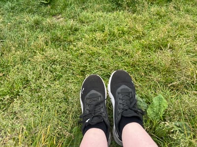 The image shows a close-up view of a person's feet, wearing black athletic shoes and black socks, standing on a grassy area. The grass is a mix of green and dried blades, with some scattered seed heads. There is a large green leaf, possibly a plantain, to the right of the left shoe. The background is out of focus, showing more of the grassy terrain. The lighting suggests an outdoor, daytime setting. The location is Tikhoretsk, Russia.