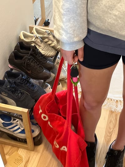 The image captures a ground-level perspective within an indoor setting, likely a home entryway or bedroom, in Aarhus, Denmark. A person is partially visible, wearing a light grey long-sleeved top over dark blue athletic shorts and black athletic shoes. Their right hand holds a set of keys with a black fob, attached to a pink and green personal safety alarm.
A bright red fabric bag, featuring white lettering with "O" and "A" visible, hangs over a wooden shoe rack. The shoe rack contains multiple pairs of athletic shoes. On the top shelf, two pairs of metallic gold Adidas shoes are neatly placed. The middle shelf displays dark athletic shoes, including one pair with a "Puma" logo partially visible. The bottom shelf holds a pair of white and blue Adidas Samba sneakers.
The flooring consists of light-toned wooden planks, with a cream-colored, fringed rug partially visible. A small piece of paper with faint handwritten text and numbers is attached to the right side of the shoe rack. The overall scene suggests the person is either preparing for or concluding an activity, given the athletic wear and accessories.