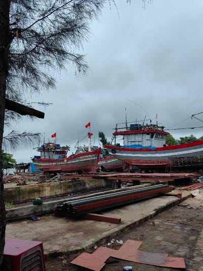 The image depicts a boat repair and maintenance yard located in Thuận An Ward, Vietnam, under an overcast sky. In the foreground, to the left, a portion of a tree trunk with sparse, needle-like foliage extends into the upper left quadrant. Below it, a stack of red plastic stools is visible next to the left edge. The ground consists of uneven concrete and dirt, with several reddish-brown, flat metal panels scattered on the right side of the foreground.

The mid-ground features a concrete structure resembling a dry dock or boat launching ramp, containing two parallel sets of metal tracks or rails. Stacked metal beams, painted red and black, lie horizontally on the concrete to the right of these tracks.

The background is dominated by three prominent traditional Vietnamese fishing vessels, dry-docked on cradles or supports.
1.  The leftmost large vessel has a dark red hull and a white upper cabin structure, flying a red Vietnamese flag from its mast.
2.  The central vessel, positioned slightly behind the first, features a dark red hull with a lighter stripe and a visible registration number "TV-696.12TS" on its bow. It also has a white upper structure and flies a red Vietnamese flag.
3.  The rightmost and largest visible vessel displays a grey and red hull with a blue tarp draped over a section of its side. It has a white upper cabin with grey detailing and mast structures.

Additional partially obscured vessels are visible further in the background. No individuals are clearly discernible actively working or interacting with the vessels or objects in the scene. The overall environment suggests an industrial area dedicated to marine vessel repair.