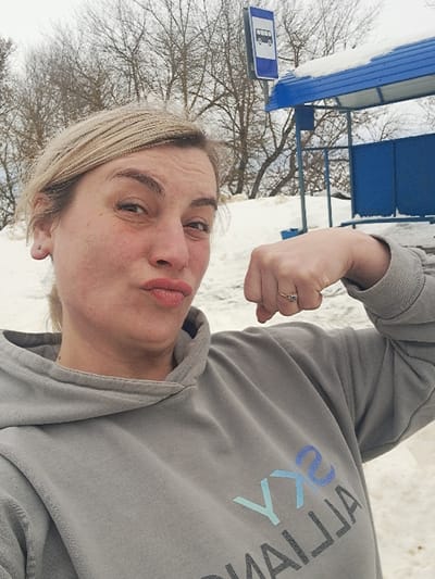 A fair-skinned female is depicted in a close-up, selfie-style photograph taken in the city of Sasovo, Russia. She has blonde hair, pulled back, and her face shows pursed lips and a slightly raised left eyebrow. She wears a grey hooded sweatshirt with the partial, inverted text "SKY ALLIAJ" visible on the chest. Her right arm is bent at the elbow, with her hand fisted and held close to her shoulder in a flexing pose, revealing a ring on her right ring finger. A small silver stud earring is present in her left ear. The background features a blue-framed bus stop shelter with a snow-covered roof, partially visible to the right. Above and slightly behind the shelter, a blue and white rectangular sign with a white bus icon clearly indicates a bus stop. The ground and distant areas are covered in snow, and bare deciduous trees are visible against an overcast sky, indicating a winter environment.