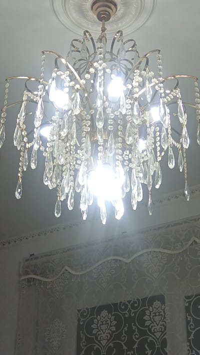 A dimly lit indoor scene features a ornate chandelier with numerous glass crystals hanging from a gold-colored metal frame. The chandelier is illuminated by several bright light bulbs, causing some areas to be overexposed. Below the chandelier, a decorative window valance with small beaded accents is visible, adorned with a swirling pattern. Further down, a patterned curtain hangs, with a darker, ornate design on the right side of the frame. The location is identified as Maqat, Kazakhstan.
