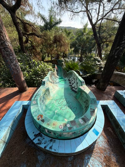 The image shows an outdoor park area in Thủy Xuân Ward, Vietnam. A defunct, turquoise, graffiti-covered fountain is the central object. It is round with a curved, raised lip and surrounded by light blue tile steps. The area is covered in dried pine needles. The background includes dense greenery, primarily trees and foliage. A section of brickwork, possibly part of a wall or other structure, is also visible. The image is taken during daylight hours.