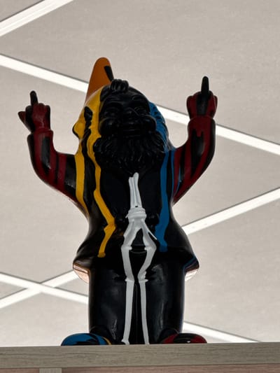A close-up photograph captures a brightly colored, stylized sculpture of a duck. The duck is painted in a vibrant palette, featuring black, yellow, blue, red, and white detailing. The sculpture is positioned against a neutral-toned wall with geometric white beams. The location is Les Arcs, France. The duck sculpture is positioned to the right of a light-colored wooden doorway. The duck extends the middle finger of its left hand, suggesting the sculpture's purpose is political, as the duck is shown giving the middle finger.