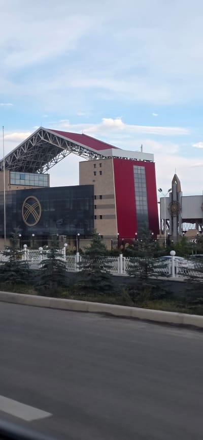 The image displays a modern architectural structure, possibly a convention center or a large public building, in Бактуу-Долоноту, Kyrgyzstan. The building features a striking design with a prominent red upper section and a dark glass facade on the lower part, which bears a large, circular emblem. A skeletal metal support structure is visible, suggesting it might be an entrance canopy or part of the building's unique architecture.
The building is bordered by a paved area, likely a road or sidewalk, and a landscaped green space with trees and white decorative fencing. Several white spherical lamps are strategically placed along the fencing. The sky above is a light blue with scattered white clouds, indicating daytime. The overall impression is one of contemporary design set against a natural backdrop, with no people or specific activities clearly depicted. The scene appears calm and orderly, likely during daylight hours. No discernible text is visible in the image.