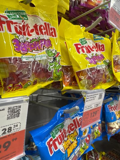 This is a close-up, eye-level shot of a grocery store display featuring packages of gummy candies. The focus is on two main types of packaging: bright yellow bags with a large, colorful "Frutella" logo and cartoon characters, and blue bags with a similar "Frutella" logo and cartoon designs. The candies themselves, in various fruit flavors, are visible through the transparent plastic packaging.
Several price tags and informational labels are attached to the display. One label, in Russian, shows a price of "128.24" and "129.99," with a discount indicated by "-11%." Another label displays "28.24" and "24." There's also a QR code visible on one of the labels. The setting appears to be a retail environment, likely a supermarket aisle, with metal shelving and other products faintly visible in the background. The lighting suggests an indoor setting, possibly during daytime hours. The overall impression is one of a well-stocked candy aisle.
Visible text includes brand names like "Frutella," descriptions such as "Мармелад жевательный" (chewy marmalade), "С фруктовым соком" (with fruit juice), "Натуральный вкус" (natural taste), and price denominations in Russian rubles. There are also references to product weight like "(Россия) 150г" (Russia, 150g). The time and date "2026 09:36" are also visible on one of the labels. No people are directly visible in the image.