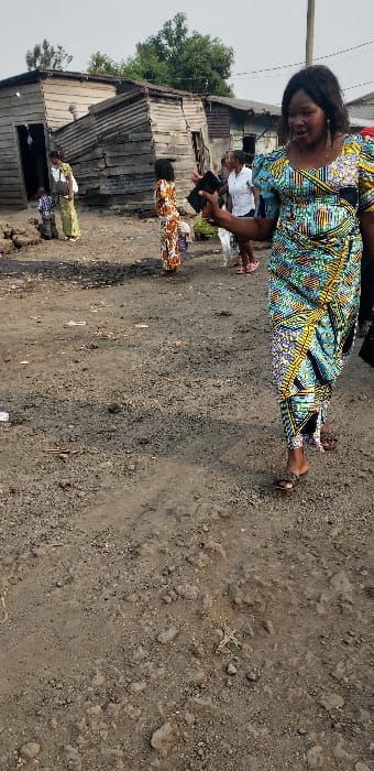 The image captures an unpaved street scene in Buzove, Democratic Republic of the Congo. The ground is composed of dry, packed dirt with scattered small rocks and dark patches. In the foreground, a woman with dark hair, wearing a vibrant, multi-colored patterned dress and brown sandals, walks towards the left, holding a dark, rectangular object in her right hand and a dark bag in her left.

In the mid-ground, several individuals are present near dilapidated structures. These buildings are constructed from weathered wooden planks and corrugated metal sheets, exhibiting uneven and worn appearances. To the left, a woman in a light-colored outfit stands near the entrance of one such wooden building, with a small child in dark clothing standing beside her. Further right in the mid-ground, another woman in a brown and orange patterned dress stands, with additional figures partially visible behind her, including one in a white shirt and red sandals. Trees and an overcast sky are visible in the background beyond the structures.