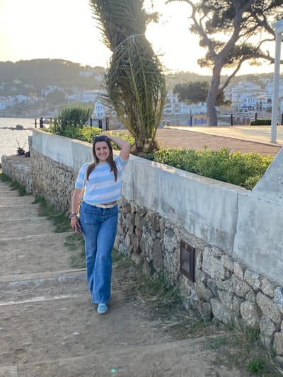 A medium shot depicts a young woman standing on an outdoor staircase, angled towards the viewer’s right. She’s wearing a blue and white striped shirt, jeans, and sneakers. She is smiling and has one hand on her head. The background suggests a seaside location in Palafrugell, Spain, evidenced by buildings on a hillside and a glimpse of the sea. A palm tree is visible on the left. The staircase is stone with vegetation alongside, and a concrete barrier separates the walkway from the scene below.