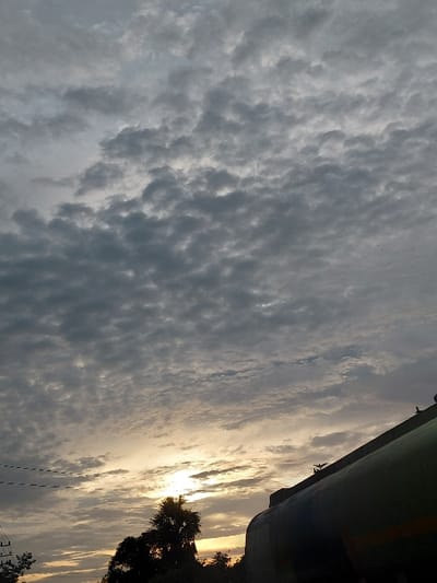 The image, captured in Lhok Mereubo, Indonesia, depicts a sky scene during either sunrise or sunset. The upper two-thirds of the frame are dominated by an overcast sky featuring dense, grey altocumulus clouds. In the lower-center, the sun, partially obscured by clouds and foreground elements, emits a bright, warm yellow-orange glow low on the horizon. The lower right quadrant features a large, dark-green, curved cylindrical structure, appearing to be a storage tank, silhouetted against the brighter sky. Two small, dark avian silhouettes are visible on the upper edge of this tank; one appears perched, the other possibly in flight. Along the lower left horizon, silhouetted trees and dense foliage are present. Several thin, dark utility lines traverse the mid-left portion of the sky.
