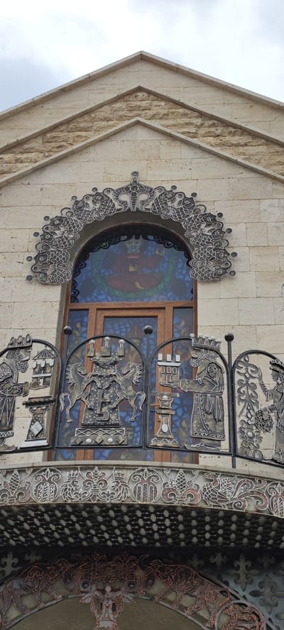 The image shows the facade of a building in Voskevaz, Armenia. The building is constructed of light-colored stone blocks. The facade features an arched window with stained glass depicting a seated figure. Above the window is ornate metalwork, resembling stylized fish or scales. Below the window, a decorative metal balcony railing displays figures and motifs, including chess pieces, horses, and human figures in elaborate attire. The balcony has intricate patterns, and below it, further metalwork with animal motifs, such as bulls, is visible. The overall aesthetic is elaborate and decorative, suggesting a building of cultural or historical significance.