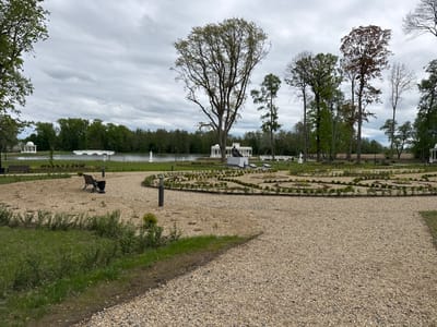 The image depicts a park scene in Добасненскі сельскі Савет, Belarus, under an overcast sky. The foreground consists of a gravel pathway leading into the park. To the left of the path is a patch of grass with scattered vegetation, and a park bench with a trash receptacle beside it.
Further into the park is a landscaped area with circular patterns of small shrubs. In the background, a lake is visible, featuring a white bridge and a white gazebo-like structure on the shore. Several statues and additional gazebo structures are also present along the lake's edge. A wooded area forms the backdrop behind the lake. Several tall trees with varying shades of green and brown foliage are interspersed throughout the park.