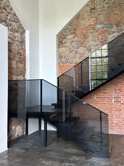 This is a photograph of a modern staircase with glass railings and dark wood treads, set against a rustic brick and stone wall. The staircase is centrally located within the frame, with one side leading upwards to the right and another section extending downwards and to the left. The brick wall, visible in the background, shows signs of age with visible mortar and varying shades of red brick and grey stones. A large window with multiple panes is visible through the glass railing on the right side of the staircase, revealing a glimpse of green foliage outside. The floor is a dark, polished concrete. The overall impression is a blend of industrial and historical aesthetics. This location is not in Ilyanskiy selskiy Sovet, Belarus.