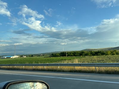 This image captures a rural landscape, likely on the outskirts of Urbnisi, Georgia, with a view from a moving vehicle. The foreground shows a highway with a white line marking the lane, and a metal guardrail separating the road from the adjacent land. Beyond the guardrail, a field of green crops stretches towards a row of trees. In the background, a cluster of buildings, including a tall communication tower and a blue-roofed structure, is visible. Further in the distance, rolling hills are dotted with wind turbines, suggesting a location where renewable energy is utilized. The sky above is a vibrant blue, adorned with scattered white clouds, and a faint sliver of the moon is discernible. The overall scene depicts a serene, open environment with a blend of agricultural and technological elements. There are no people visible in the image, and no discernible actions or interactions are taking place.