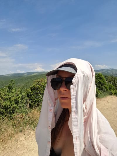 A woman wearing sunglasses and a light pink shirt pulled over her head, possibly for sun protection, is taking a selfie in a natural landscape. The background shows rolling green hills covered in dense trees, with a dirt path in the foreground. The sky is a clear blue with some wispy clouds. The location is identified as Bol'shoi Utrish, Russia.
