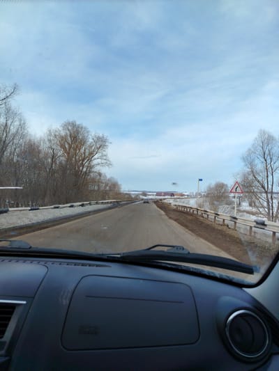 The image presents a forward-facing view from inside a vehicle, looking out through the windshield at a road scene in a rural or semi-rural area within the city of Krasnoyarsk, Russia. The vehicle's dark-colored dashboard occupies the lower portion of the frame, showing air vents on the left and a circular vent with a chrome ring on the far right, with a windshield wiper blade visible at the base of the windshield.
The two-lane asphalt road, appearing damp and bordered by muddy shoulders with patches of snow, extends into the distance, gently curving to the right. A metal guardrail, painted white with black striped sections, lines the right side of the road, with snow and dirt visible beneath it. To the left of the road, a dense line of bare deciduous trees and shrubs forms a natural barrier, with snow accumulated on the ground. On the right, beyond the guardrail, similar bare trees and shrubs are scattered across snow-covered fields.
Two road signs are visible on the right: a blue rectangular sign displaying the number "11" in white, and a triangular warning sign with a red border and a black right-curving arrow, indicating a curve ahead. In the far distance on the road, a dark-colored vehicle is observed traveling in the same direction. The background reveals a flat, snow-covered landscape with several low-rise buildings, identifiable by their reddish roofs. The sky is predominantly light blue with sparse, wispy white clouds.