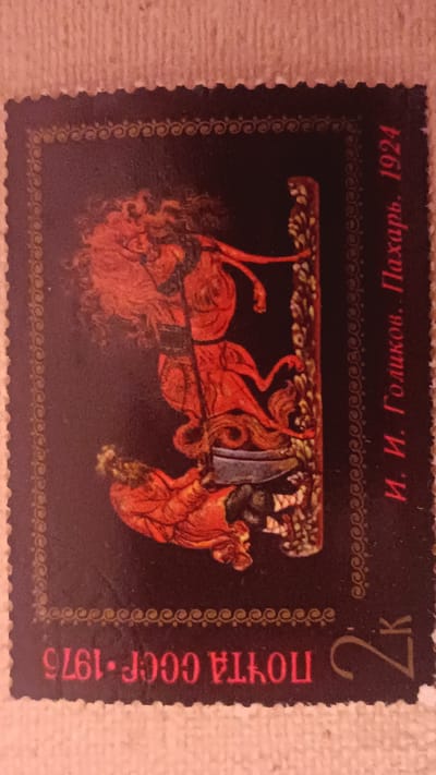 This is a close-up, angled shot of a postage stamp. The stamp features a dark background with a decorative border. The central image depicts a scene from a folk tale or myth, rendered in a bright, stylized manner with dominant red and orange colors. Two figures are prominent. The main figure, positioned on the right and slightly elevated, appears to be a female character with long, flowing red hair, dressed in ornate red and brown attire. She is in a dynamic pose, possibly fighting or engaged in a ritual, holding a long, dark object that extends downwards. Below her, to the left, is another figure, also in red clothing, lying on the ground in a supine position, possibly defeated or injured. The figures are depicted with bold outlines and simplified forms, characteristic of folk art. There is a vertical, decorative element on the right side of the stamp, resembling a tree or column adorned with patterned leaves.

Visible text on the stamp includes Cyrillic script. Along the right edge, oriented vertically, is "И. И. Голиков. Пахарь. 1924", which translates to "I. I. Golikov. Ploughman. 1924." Along the bottom edge, also in Cyrillic and oriented horizontally but reversed, is "ПОЧТА СССР 1975", indicating "POSTAGE USSR 1975." In the bottom right corner, there is a prominent numeral "2" followed by a Cyrillic letter "к", likely indicating a denomination of 2 kopecks. The overall impression is one of dramatic action and vibrant color, suggesting a narrative scene from Russian folklore or history. The stamp was issued in 1975, commemorating a work from 1924 by artist I. I. Golikov. The location context of Onaldy, Turkmenistan, is not visually represented on the stamp itself but is provided as external information.