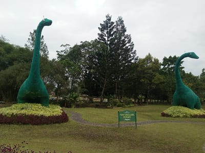 A park scene featuring two large, green dinosaur topiary sculptures resembling Brontosauruses. Each stands atop a bed of vibrant flowers. A sign reads "Twin Dinosaurs Topiary" sits between them. Green grass covers the ground, interspersed with a paved walkway. A variety of trees fill the background under an overcast sky.
