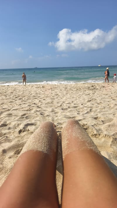 The image depicts a beach scene. In the foreground, a person's legs covered in sand are visible, suggesting they are lying on the beach. The middle ground features a sandy beach with several people in the distance. One individual is standing near the shoreline in what appears to be swimwear. Another person, possibly a woman, is seen wearing a hat and a dark swimsuit with a child beside her. The background consists of the ocean and a partly cloudy blue sky. A boat can be seen on the water in the distance. The location is a beach, but not located in the Jiyang District, China.