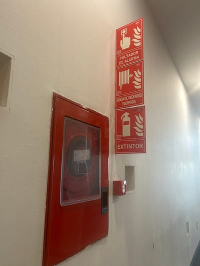 This image captures a section of an interior corridor or hallway in Leganés, Spain, featuring mounted fire safety equipment. A red fire hose cabinet, partially recessed into an off-white wall, occupies the left-center of the frame. Its transparent front panel allows visibility of a coiled red fire hose and nozzle housed within. A black handle is integrated into the right side of the cabinet, and the number "36" is visible on its upper right edge.

Vertically aligned above and slightly to the right of the fire cabinet are three rectangular red fire safety signs with white iconography and text. The top sign displays a hand pressing a button, labeled "PULSADOR DE ALARMA" (Alarm Button). The middle sign features a fire hose reel, labeled "BOCA DE INCENDIO EQUIPADA" (Equipped Fire Hydrant/Hose). The bottom sign shows a fire extinguisher, labeled "EXTINTOR" (Fire Extinguisher). Each sign includes a small "SPV" logo and a flame symbol.

Below the lowest sign and to the right of the fire cabinet, a red manual fire alarm call point is surface-mounted on the wall, positioned next to a small, empty rectangular wall niche. Another, larger empty rectangular niche is cut into the wall to the left of the fire cabinet. A faint vertical hairline crack extends downwards from the top edge of the image, running to the left of the fire cabinet. The environment is illuminated by ambient lighting, creating a gradual gradient of light on the right-hand wall. No individuals, actions, or interactions are depicted.